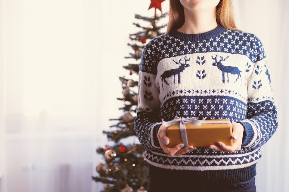 personne portant un pull de noël et tenant un cadeau devant un sapin